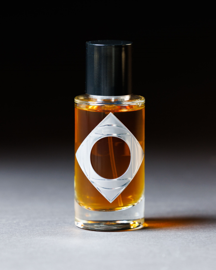 A small glass bottle filled with amber liquid, featuring a black cap and a silver geometric label with a circular cutout, evokes the elegance of luxury nameplates on a neutral surface against a dark background.