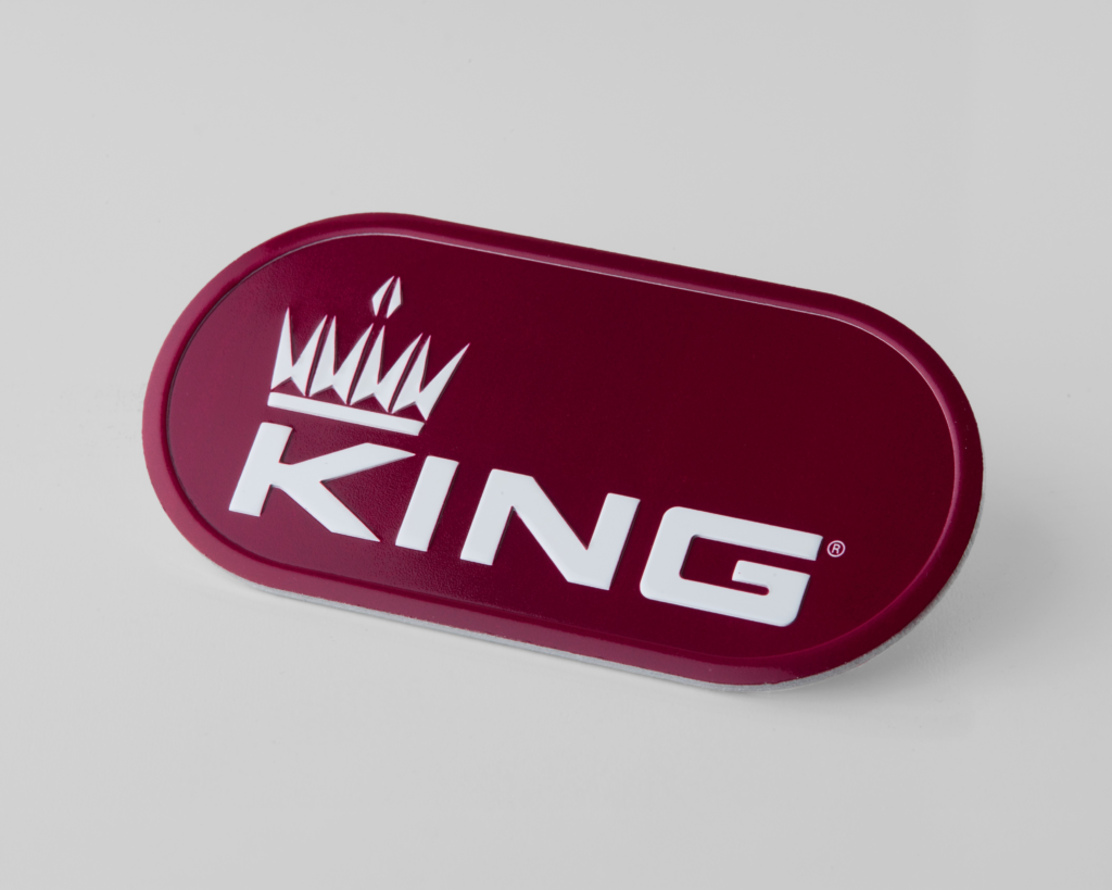 A maroon oval badge with a white crown graphic above the word KING in bold, white letters, set against a plain light gray background, reminiscent of classic equipment nameplates.